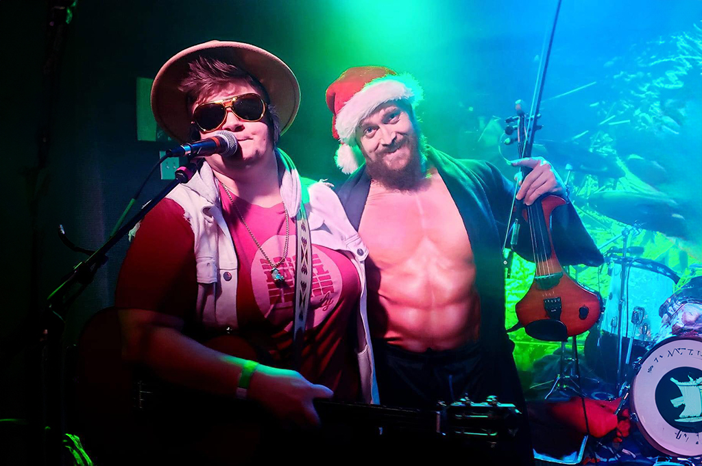 Two performers on stage; one wearing a Santa hat and a vest, and the other in a red sleeveless shirt and a white hat, both with sunglasses on. The one in the Santa hat is singing into a microphone while the other holds a violin, with stage lights giving a green hue to the background.