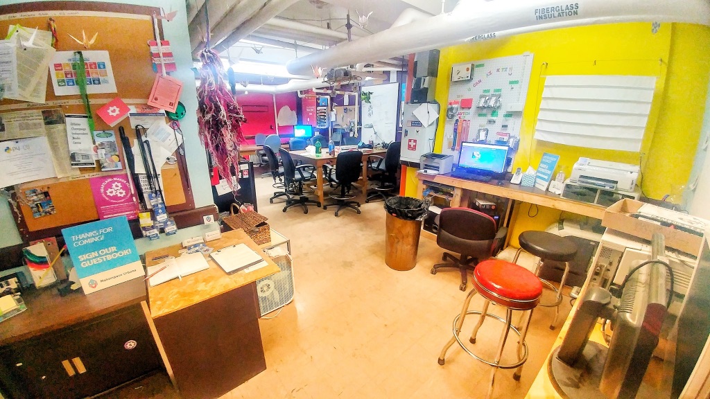 A very full room with computer equipment, craft supplies, and many desks and chairs. There is a cluttered cork board on the left, a bright yellow wall on the left, and a pink wall in the back. There are pipes and duct work in the ceiling and bright overhead lights.