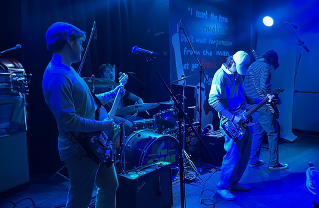 A band is illuminated by soft blue stage lighting, enhancing the intimate atmosphere of the venue. The guitarist in the foreground on the left stands with a black electric guitar, and his gaze seems to be directed towards the camera, giving an inclusive feel to the viewer. The drummer, centered in the background, is actively engaged in playing, with a visible expression of enjoyment. To the right, another musician's back is to the camera, showcasing a white bass guitar, while a fourth band member stands facing away, seemingly adjusting their equipment.