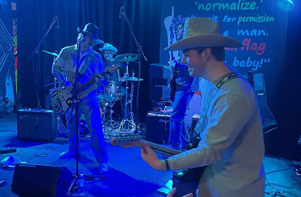 In the first image, a trio of musicians is performing on a stage with a blue ambiance. The guitarist in the foreground is wearing a wide-brimmed hat, and a musician in the background is partially visible behind the drum set, also sporting a hat. The stage features an array of microphones and cables.