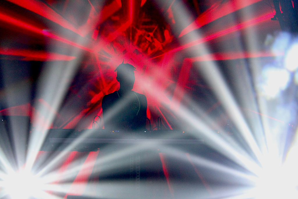 A DJ is at the center of converging red beams of light that create an explosive, almost supernova-like effect, amplifying the sense of energy and movement associated with the EDM performance.