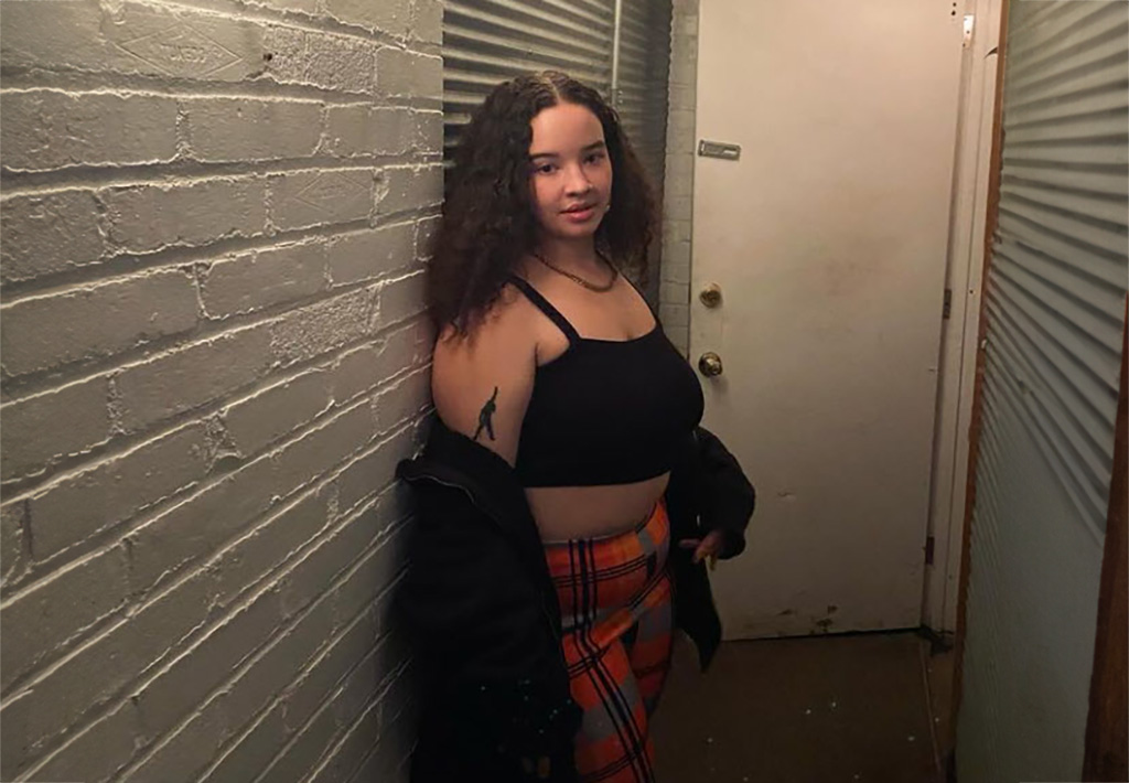 A young woman in a black halter top and plaid pants stands in a narrow hallway that looks like it's backstage at a music venue.