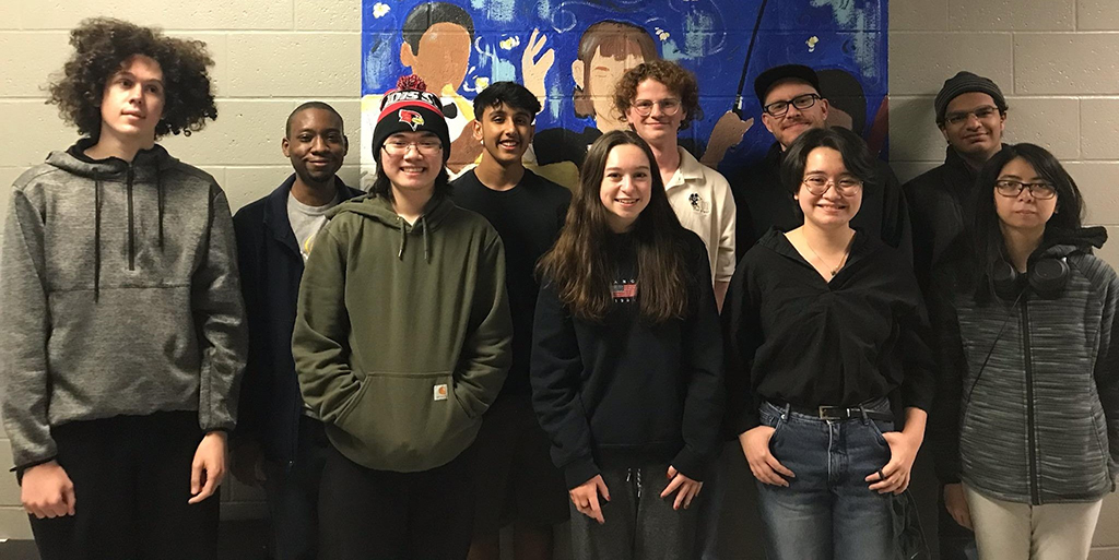 A diverse group of students stand in front of a colorful mural featuring a figure raising their hand. The students display a casual yet coordinated style, with several wearing hoodies and comfortable tops, and one sporting a sports team cap. They exhibit a variety of expressions from smiles to neutral looks, indicating a relaxed and informal atmosphere. Their attire ranges from a dark jacket and light-colored tops to a black blouse and grey zippered jacket. Headphones are worn by one individual, suggesting a connection to music or technology. The group's composition and the mural behind them convey a sense of creativity and community, typical of a collaborative educational or artistic setting.