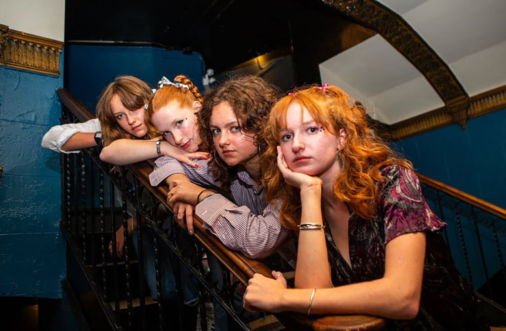Four individuals are posed in a line leaning on a stairway railing. Starting from the left, the first person is wearing a light blue button-up shirt with sleeves rolled up to the elbows. The second individual has on a white shirt with thin black stripes, a choker-style necklace, and a forearm tattoo. The third person dons a brown striped shirt and multiple bracelets on their wrist, while the last individual on the right is in a patterned dark purple blouse with puffy sleeves. They all have curly hair, varying in shades from blonde to auburn, and are adorned with hair accessories such as clips and scrunchies. The staircase behind them has dark blue walls, and the railing is a dark wood tone with ornate metal balusters.
