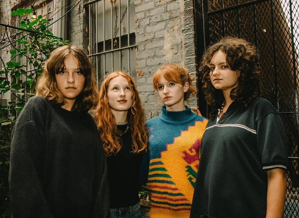 Four individuals stand against a brick wall with a barred window and green foliage around them. From left to right: the first person wears a dark sweater; the second, with long red hair, wears a blue shirt; the third has a multicolored sweater; and the fourth wears a dark top with a white collar and trim.