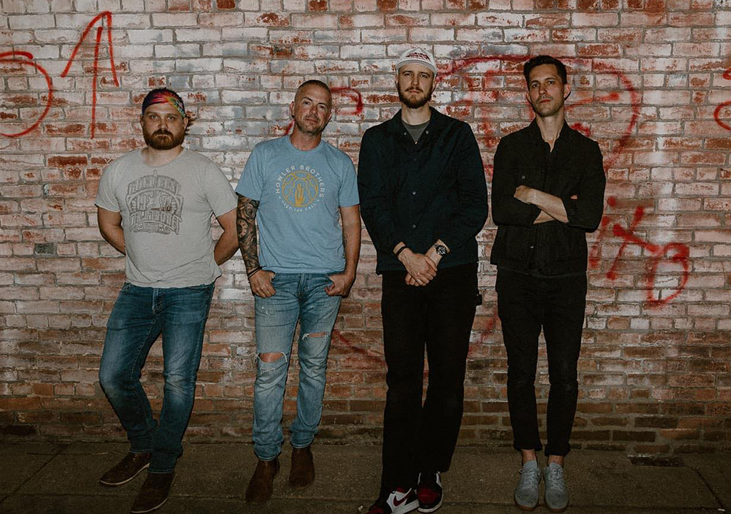 The image features four men standing against a brick wall with graffiti. From left to right: the first man has a full beard, wearing a gray T-shirt and jeans, the second man is wearing a light blue T-shirt with distressed jeans, the third man is in a black beanie and navy shirt, and the fourth man is in a black shirt with arms crossed. They all wear casual footwear. The setting suggests an urban or industrial area, and their style implies they could be a band or a group associated with a creative endeavor. The casual poses and attire, along with the graffiti background, give the image a relaxed, yet edgy vibe.