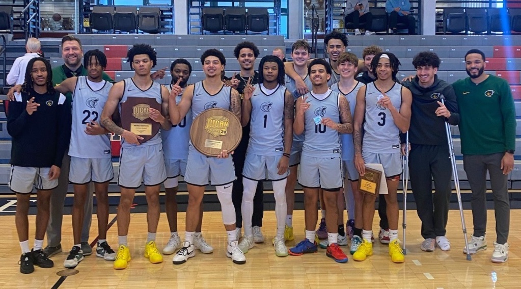 A group of young men wearing gray jerseys with green writing, stand together holding the Region 24 Championship trophy. They are standing on a basketball court.
