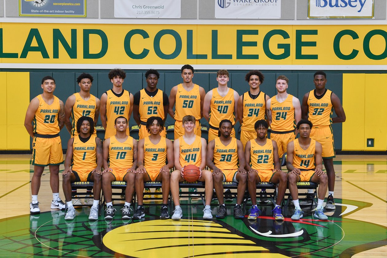 The Parkland Men's basketball team standing and sitting in the Parkland gym in two rows. They are wearing yellow jersey's with green letters and numbers on their jerseys. the player in the center front row is holding an orange basketball.