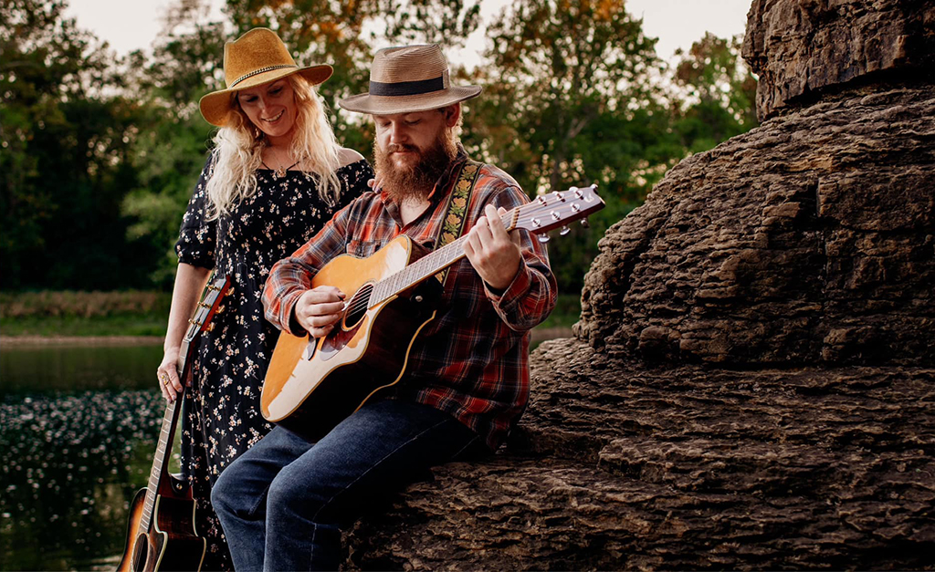 Two musicians outdoors, one in a floral dress and a wide-brimmed hat, and the other in a checkered shirt and a straw hat. They are performing together, one playing a guitar, beside a large tree trunk, with a serene water body in the background.