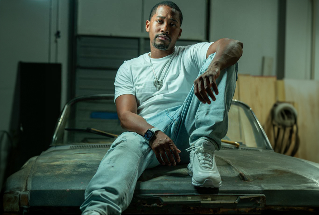 A man in a white shirt and light blue jeans sits on the hood of a car in a garage.