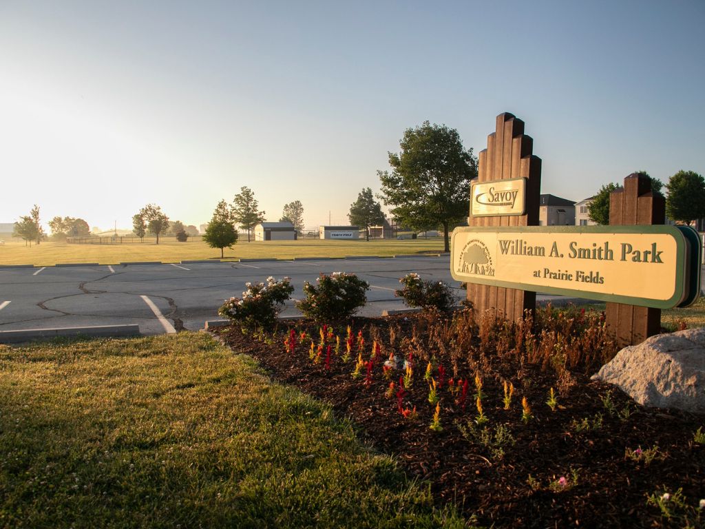 A sunrise over a big open field and a park sign that says William A. Smith Park with red flowers planted around it. There is a parking lot behind the sign. 