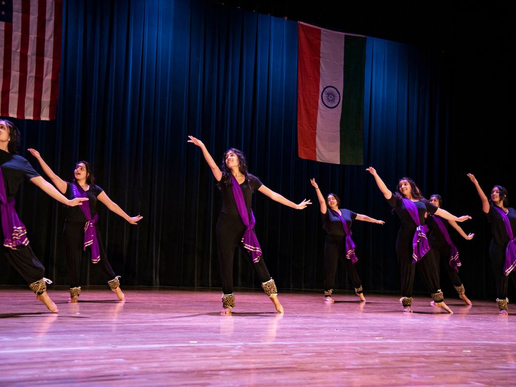 A group of women perform onstage. They wear black pants and t-shirts, with a purple sash going accross one shoulder, they have golden bells on their ankles and are in the same dance position, reaching up with only one foot on the ground. 