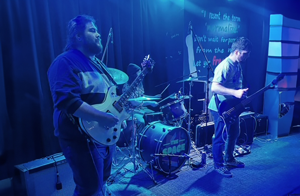 In the first image, under a blue-hued light, a trio of musicians engages with their instruments on a stage adorned with a drum set labeled "The Space". The musician on the left is playing a white electric guitar, while the one on the right is focused on a dark-colored bass guitar. Both are standing as they perform. Center stage, behind them, is a drum kit awaiting the drummer. The backdrop features a handwritten chalkboard with musings about normality and passion.