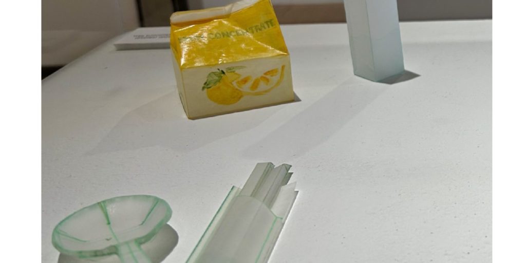 Stark photo of paper sculpture which includes a yellow fruit juice box and a spoon and chopsticks made of of thin clear vellum paper.