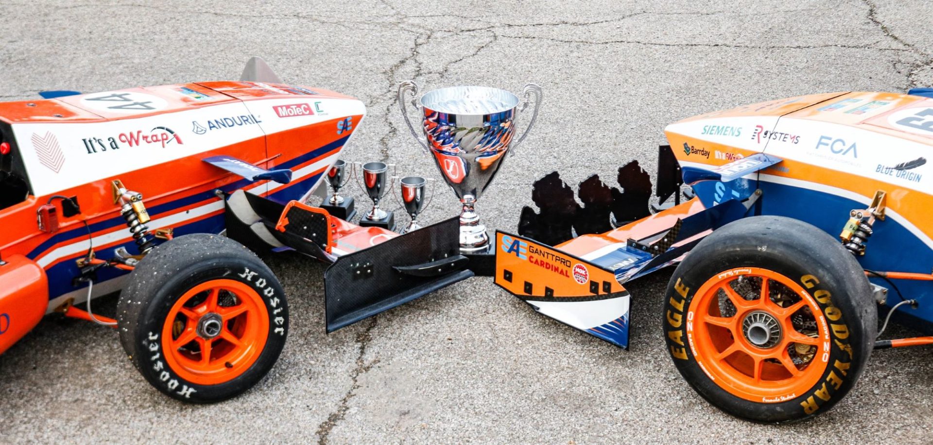 Two motorsports vehicles are nose to nose on a slab of pavement, with a trophy placed between them.