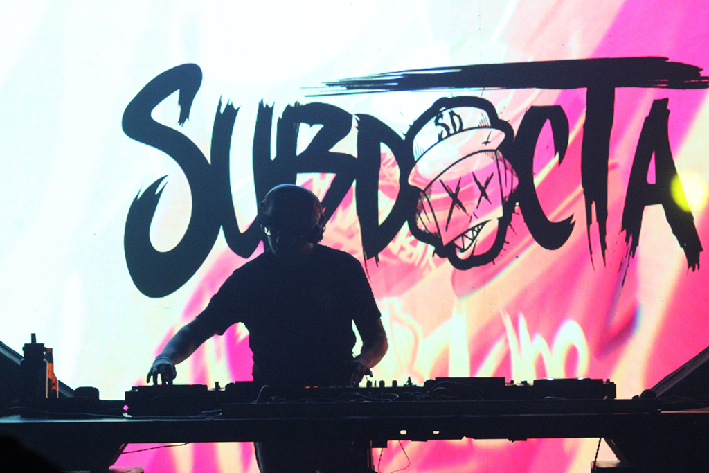 A DJ's silhouette set against a vivid, pink and black background with a prominent, stylized text that reads "SubDocta." The text and imagery on the screen have a graffiti-like style, suggesting a blend of urban art and electronic music. The performer is engaged with their setup, contributing to the lively visual and auditory experience for the audience.