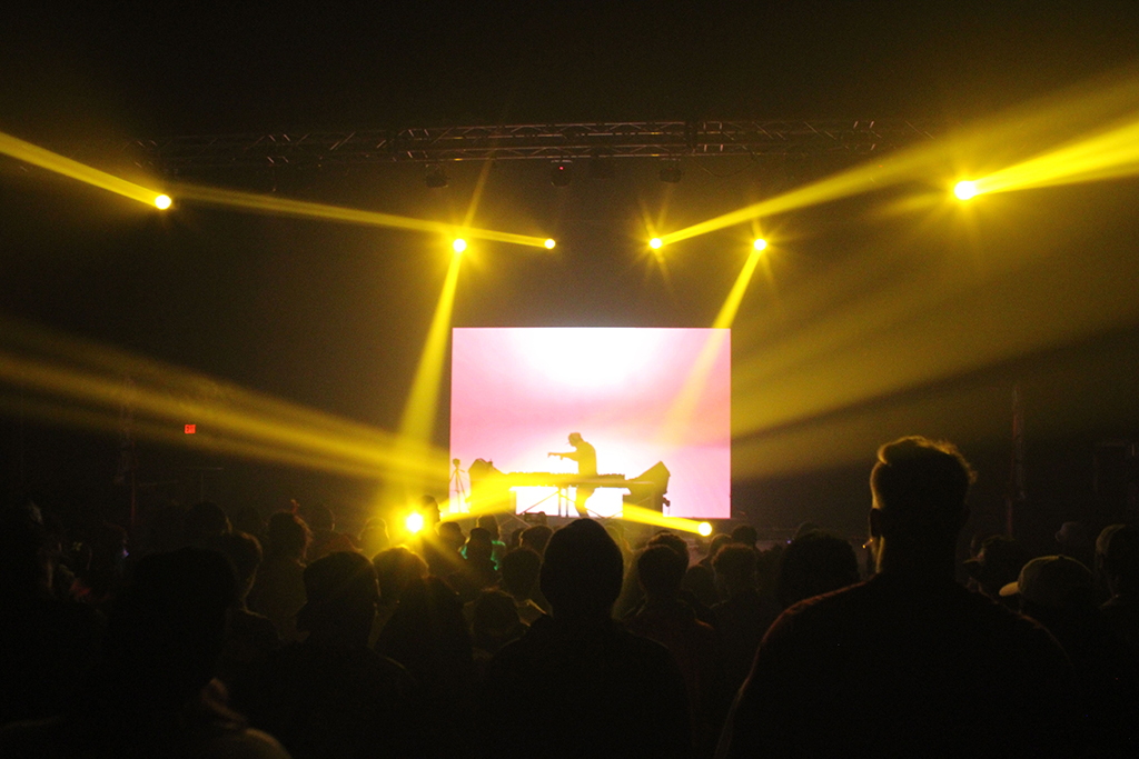 A DJ performance on stage bathed in a warm, golden glow from the lighting above. Beams of light shine outwards over the audience, converging at the artist in the middle. The stage is backlit with a soft, pink hue that graduates into a deep yellow at the center, giving a sunset-like effect. The crowd is silhouetted against the lights, creating an intimate atmosphere focused on the performer.