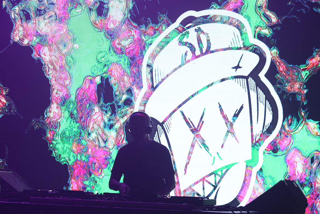 A DJ performing with a backdrop displaying a distinctive, stylized logo that appears to be a character's head. The backdrop features a psychedelic swirl of colors, lending a surreal look to the scene. The DJ is wearing a dark cap, further concentrating on their craft as they manipulate the audio equipment.