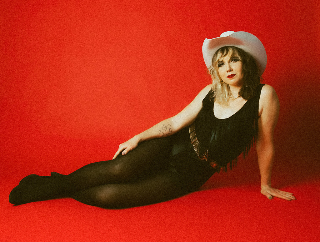 An individual lounging against a red background, dressed in black with sheer tights, a fringed top, and a white cowboy hat, exuding a vintage, country music vibe.