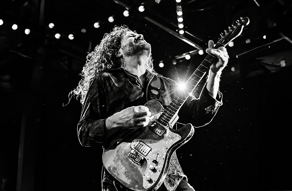 A guitarist is captured in a monochrome tone, hair tousled and mid-motion, evoking a sense of energy and passion. The spotlight shines down, illuminating the artist and the guitar, contrasting the dark background.
