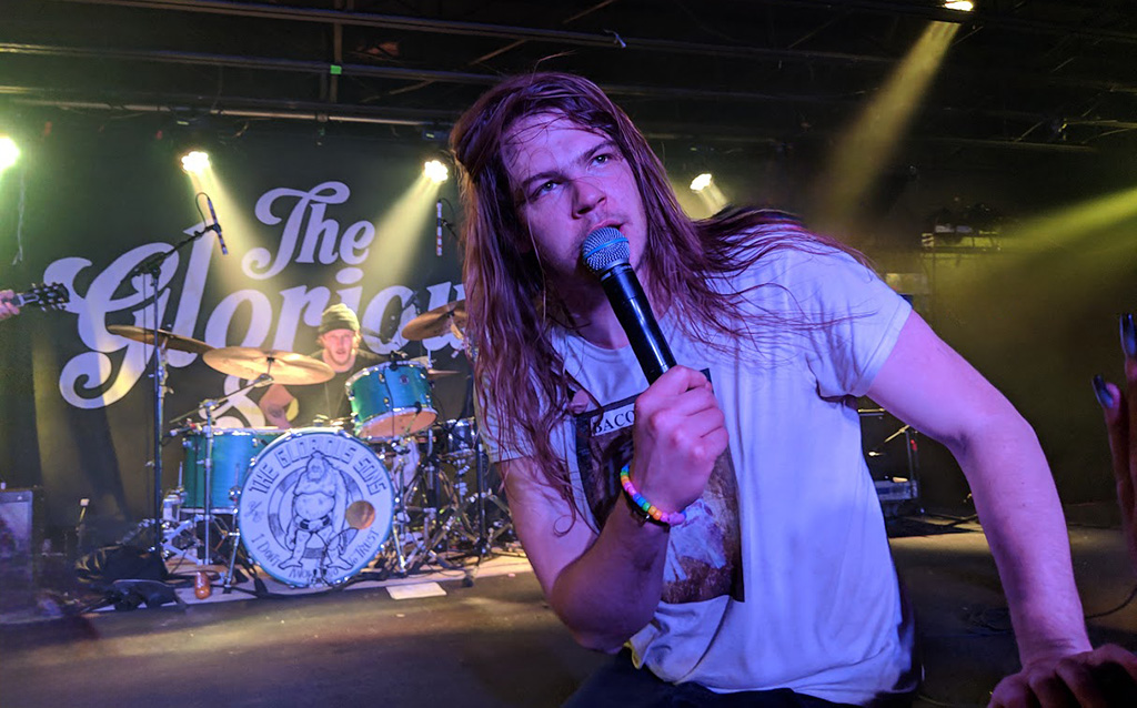 A singer is at the forefront of a lit stage. In the background you can see the drummer, and behind the drummer is a large banner that reads "The Glorious Sons".