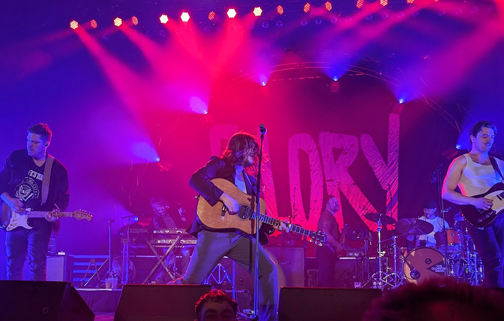 6 band members on a stage lighted in red and blue with a banner that reads "Glory" at the back of the stage.