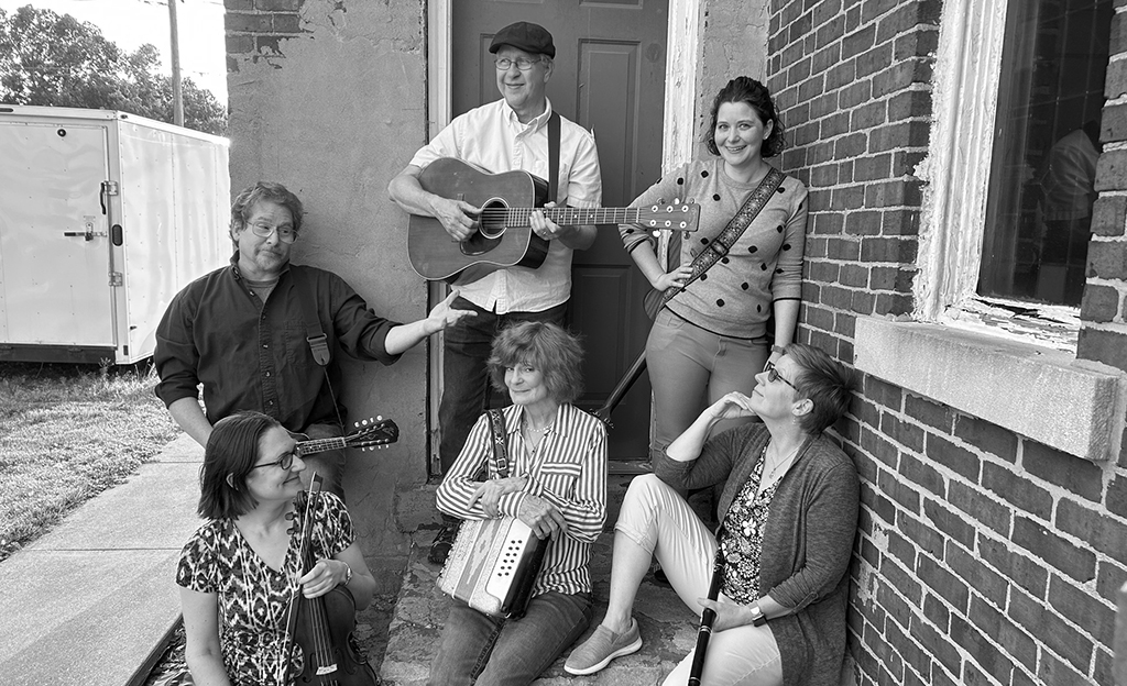 The black and white image shows a group of five musicians posing casually by an old brick building. Two are seated; one holds a violin, and the other has an accordion on her lap. Three are standing; the central figure holds a guitar, a woman to the right carries a stringed instrument, possibly a mandolin, across her body, and another is leaning against the wall, looking upwards. They all appear relaxed and content, suggesting a folksy or acoustic band vibe. The setting with an old trailer in the background and a weathered door to the left adds a rustic charm to the scene.