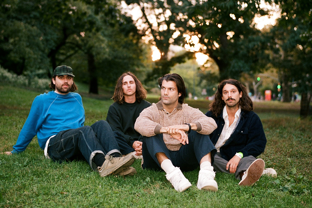 Four band members seated on a grassy knoll in a park setting, with a cityscape in the distance. The lighting suggests a sunset ambiance, and they are casually dressed, projecting a relaxed atmosphere.