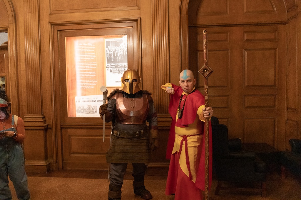 two people dressed as characters face the camera in character. One is wearing a brown outfit with a bronze chest plate and helmet and is holding a hammer. The man next to him is wearing yellow and red robes, holding a golden staff, and has a blue arrow on his bald head. 