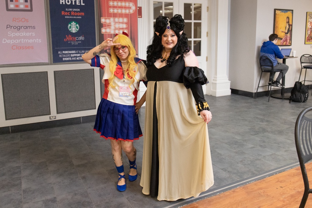 Two women stand on a gray tile floor in front of a white wall with posters. One is wearing a blue, white and red sailor top and a blue and red skirt with blue ballet shoes. She is making a peace sign. the other woman has elf ears and a long black dress with a cream overlay over her skirt. She has a silver metal piece hanging down on her forehead. 