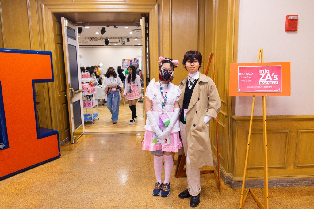 Two people stand next to each other in the doorway to the ballroom. They are dressed in costume. The woman on the left is wearing a pink and white ruffle dress with pink and white knee-high socks, and pink ears. The man on the right is wearing long khaki pants a black best, white shirt, and trench coat.  