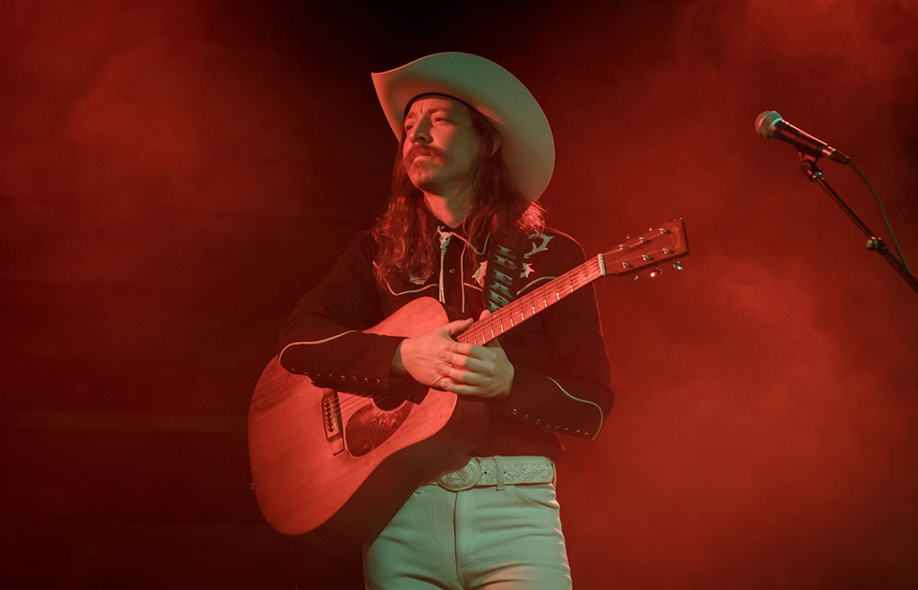 A musician is spotlighted on a dimly lit stage, wearing a cowboy hat and western shirt similar to the first image, focused intently on playing the guitar. The red stage lights cast a dramatic effect on the performance.