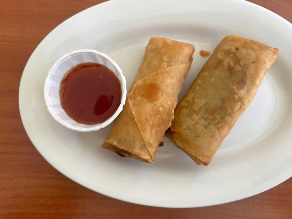 Two egg rolls on a white plate beside a paper cup of red sauce.