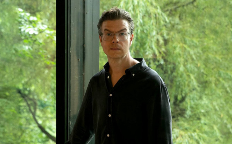 A white man with short brown hair and glasses, standing in front of a large window. There are green trees in the background.