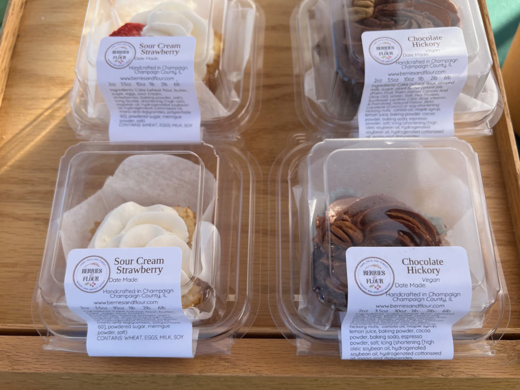 Four individually packaged small cakes by Berries & Flour are on a wooden tray.