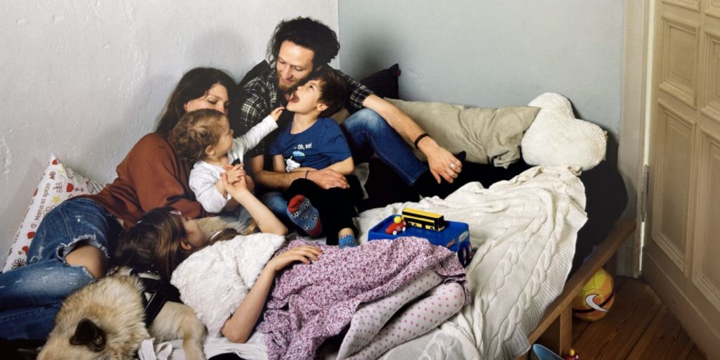 Photo of a family with a father and mother holding three young children on a bed strewn with toys. The children are touching each other and there is a blue box holding a toy school bus on the bed with a yellow and pink soccer ball under the bed.