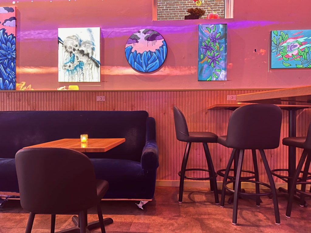 Inside Gallery Art Bar, there is a blue velvet couch and wooden table with a plush black chair. Art is on the walls.