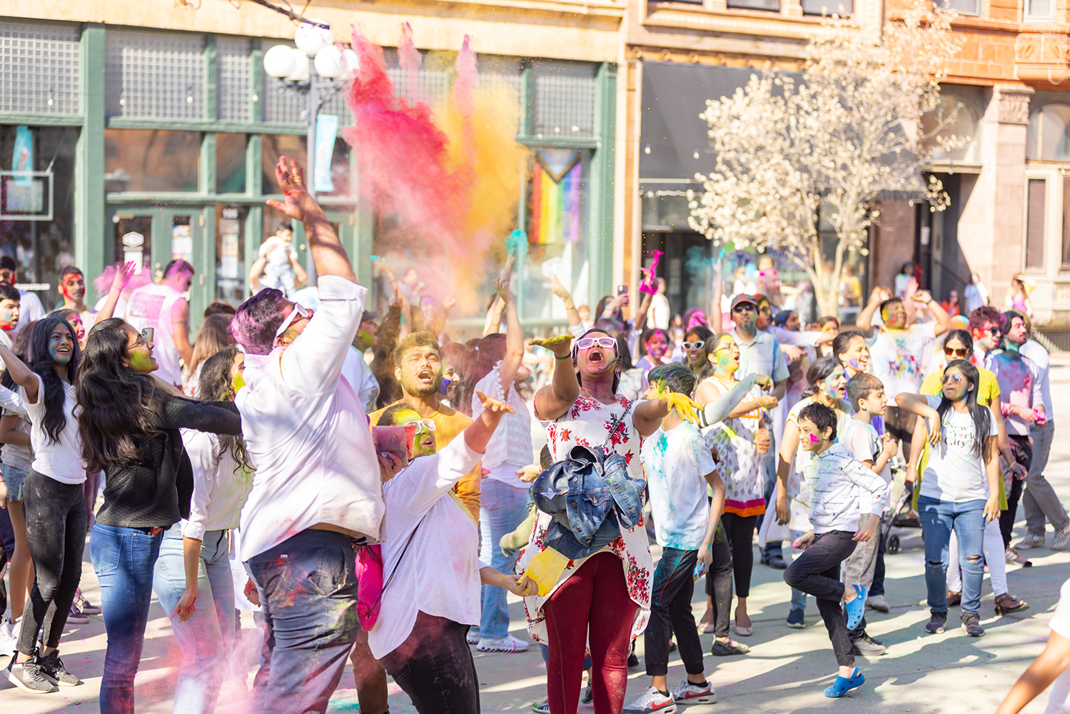 A gathering of people on a downtown street tossing color powder into the the air to celebrate Holi
