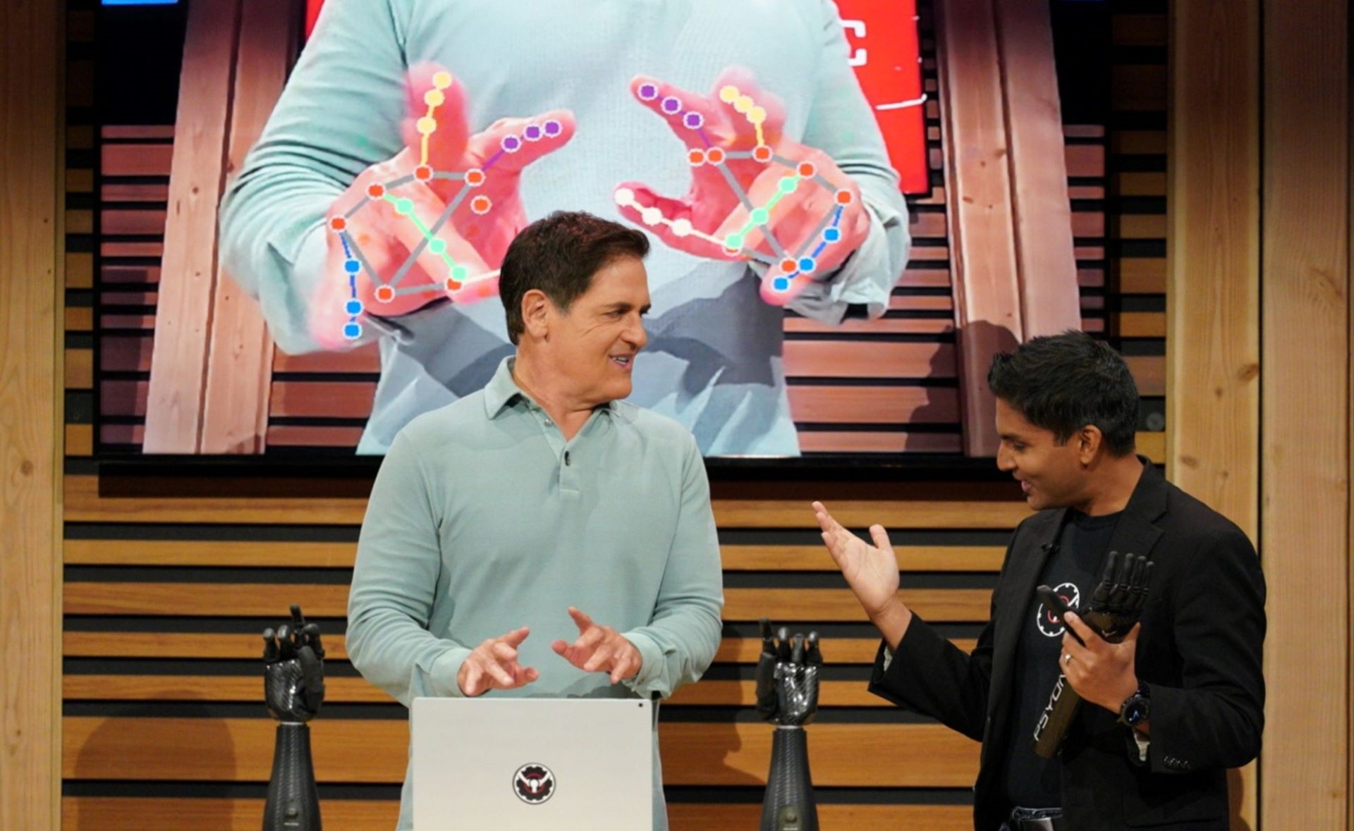 Shark Tank's Mark Cuban is standing behind a desk displaying two bionic hands, while Aadeel Akhtar stands nearby with his hands out.