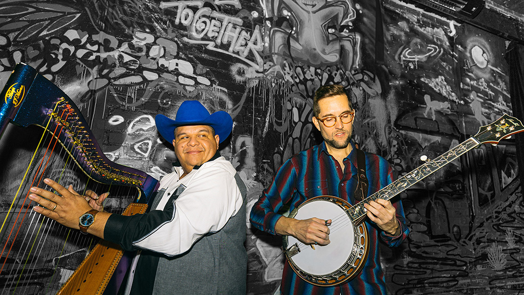 Two musicians are captured in a vibrant, graffiti-covered environment, adding a lively, urban feel to the scene. The musician on the left is playing a large, colorful harp and is dressed in a cowboy hat, light jacket, and dark shirt, which contrasts with the bright strings of his instrument. The musician on the right is playing a banjo, wearing a plaid shirt and glasses, which complements the artistic and eclectic atmosphere suggested by the graffiti background. The overall setting emphasizes a blend of traditional musical elements with a contemporary, artistic urban backdrop.