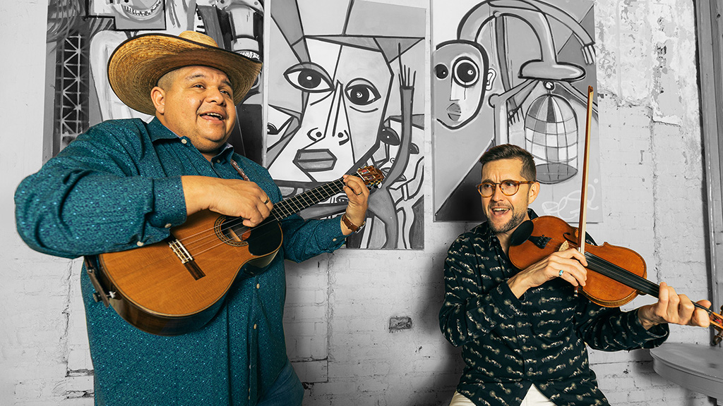 Two musicians are performing against a backdrop of abstract black and white art. The musician on the left plays a small, four-stringed instrument and wears a wide-brimmed straw hat, a teal blue shirt with a sparkle pattern, and dark pants. The musician on the right plays a violin and is dressed in a green sweater with a leaf pattern, black glasses, and dark pants.