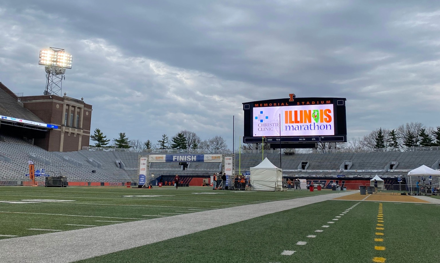 The finish line of the Christie Clinic Illinois Marathon in Memorial Stadium at the University of Illinois. The finish line is in the distance with a big blue with white text banner that reads FINISH. There is a large jumbotron screen behind that says Christie Clinic and Illinois Marathon. There stadium is empty.