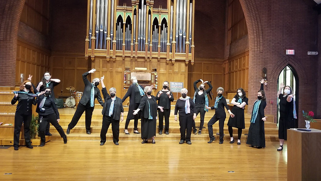a group of people standing on the steps inside what appears to be a church, with a large pipe organ in the background. They are striking various poses, some with arms stretched out or raised, evoking a sense of movement or dance. The setting suggests that this could be a choir or musical ensemble in a playful moment, contrasting the formality typically associated with such a venue.