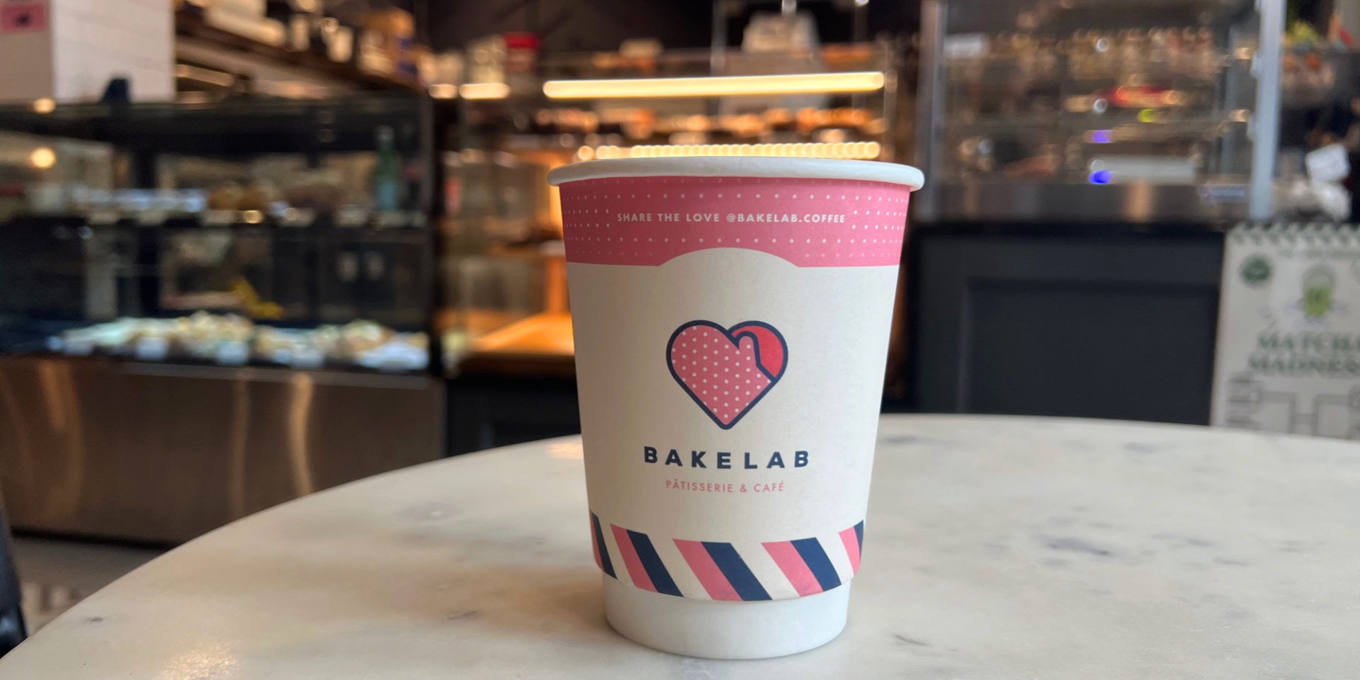 A white paper cup with the branding of BakeLab sits on a white marble table inside the coffeeshop.