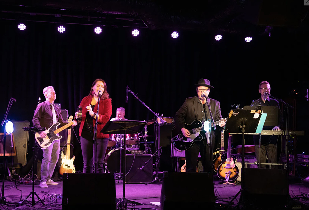 5 band members performing onstage including a male bass player, male drummer, female singer, male with a guitar that's singing and a male in the back playing keyboards.