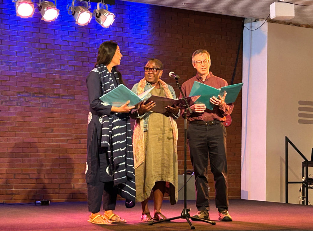 On a stage with a brick wall background, an Indian woman, a Black woman, and a white man stand in front of a microphone reading from three-ring notebooks.