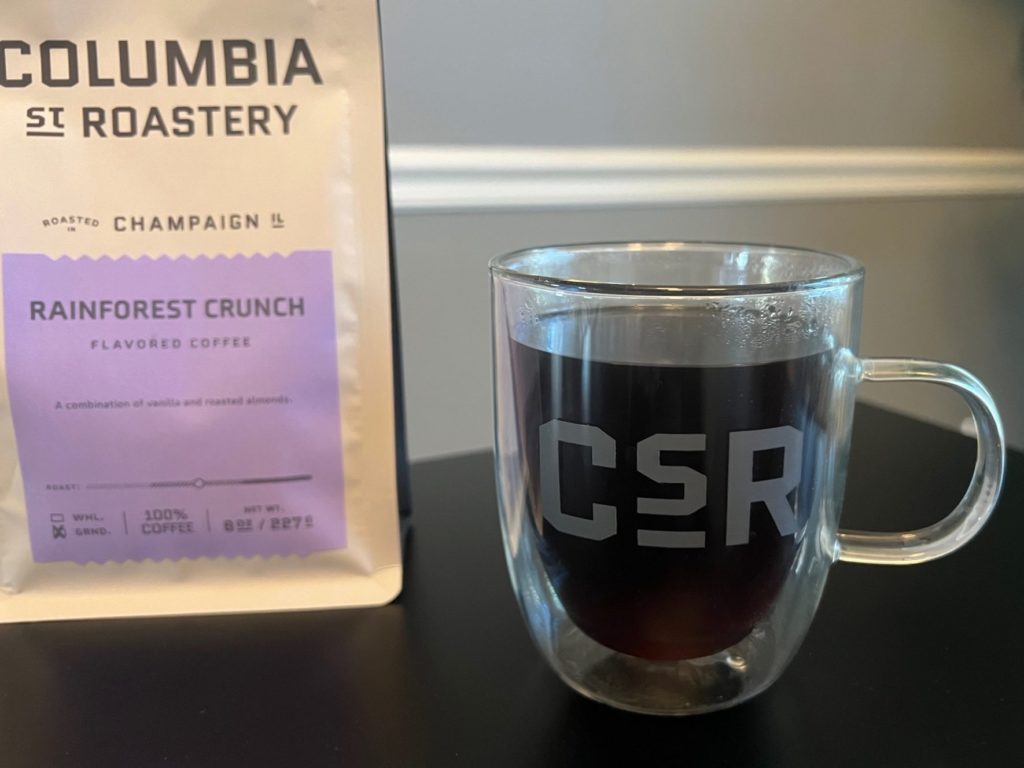 A glass mug that reads CSR with black coffee beside a bag of Columbia Street Roastery called Rainforest Crunch on a black table.