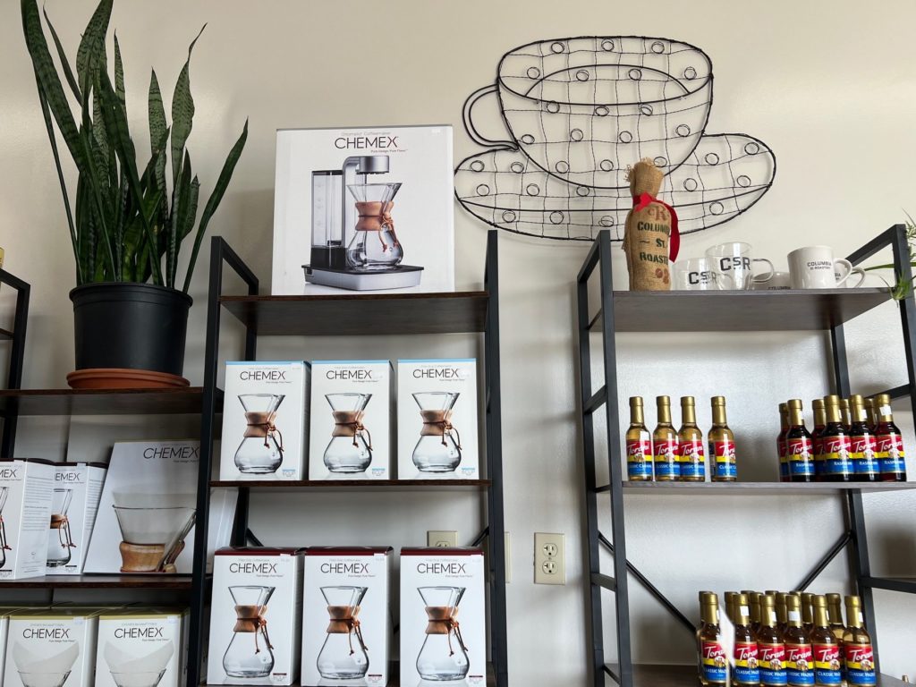 Shelves of Chemex and coffee syrups along a beige wall with a wire coffee cup decoration.