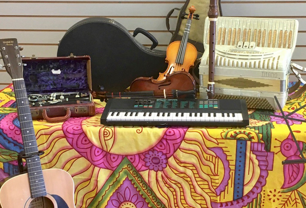 On a table with a bright patterned tablecloth sites a clarinet in its open case, a guitar is leaning on the table, there is a guitar case in back, there are two violins, an electronic keyboard and an all white accordian.