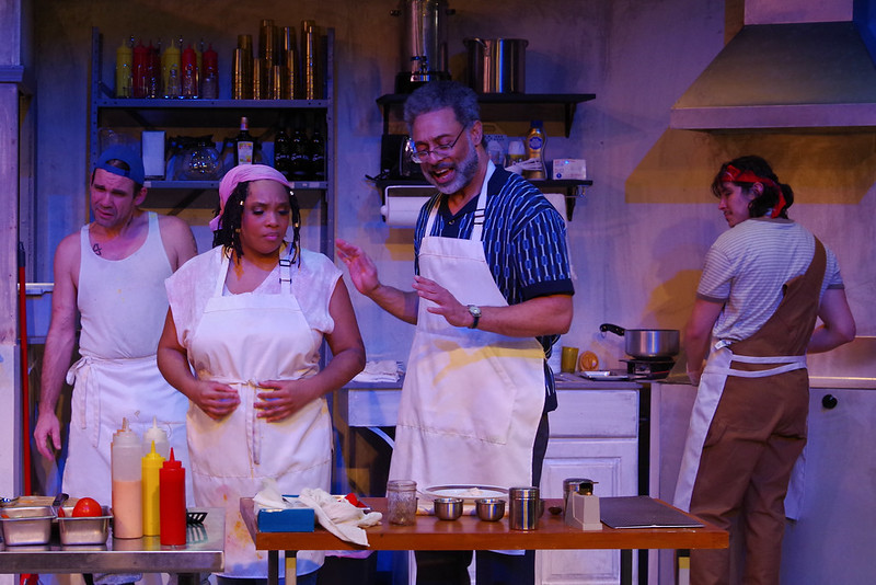 Four actors are on stage with a set that looks like the kitchen of a restaurant. A Black woman and a Black man are talking in the foreground while two white male actors are helping with food preparation.